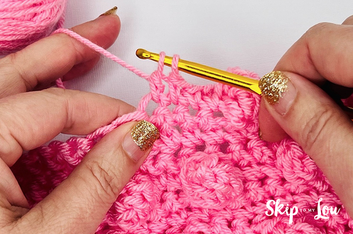 Two hands holding a crochet hook and working with pink yarn on the sixth step of the popcorn stitch.