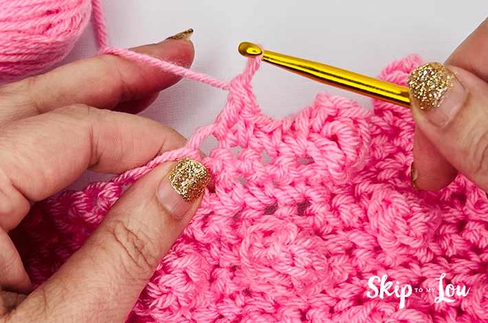 Two hands holding a crochet hook and working with pink yarn on the seventh step of the popcorn stitch.
