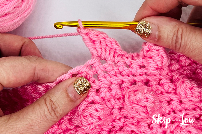 Two hands holding a crochet hook and working with pink yarn on the ninth step of the popcorn stitch.