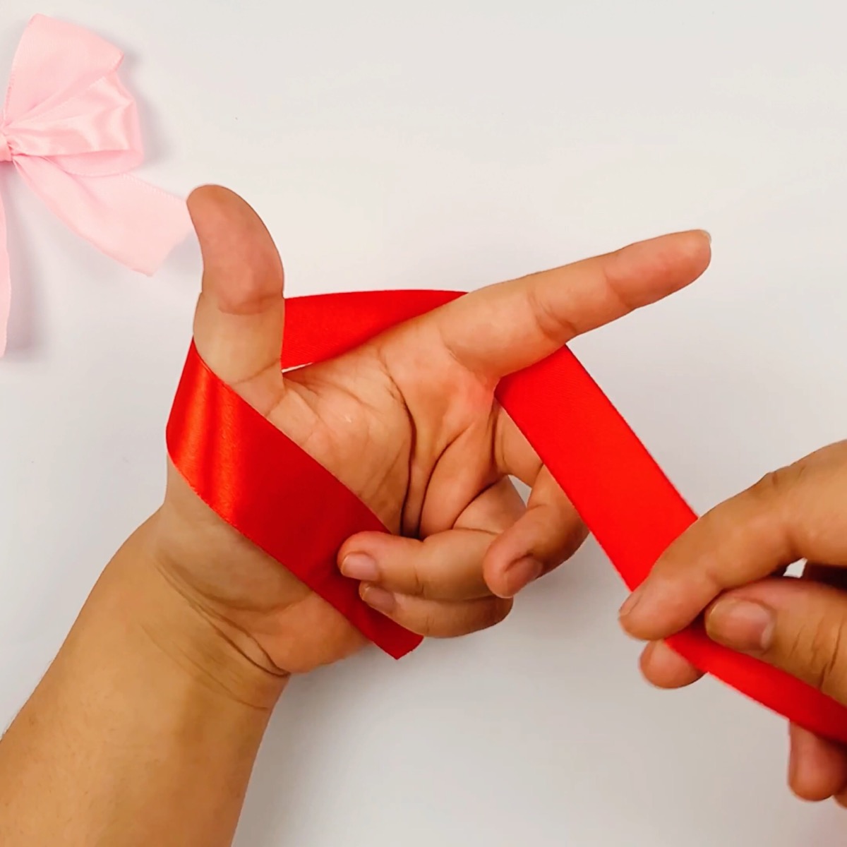 Step 1 of the ribbon bow making process showing a hand with the thumb and forefinger formed in an L-shape and the ribbon in the starting position, placed behind both of them.