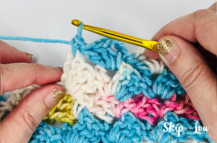 Hands holding a crochet needle crocheting the first part of the traditional tulip stitch with the needle pulling the yarn upwards after hooking it. 