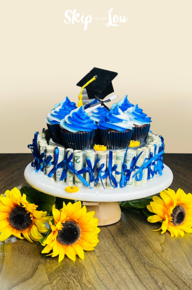 image of a "money cake" with dollar bills tied with blue ribbon and chocolate cupcakes with blue-and-white-colored frosting on top, and a graduation cap topper.