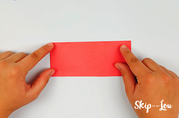 Two hands folding a red rectangular piece of paper in half. 