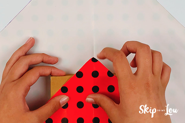 Two hands folding the bottom triangle of the wrapping paper over the top of the box.