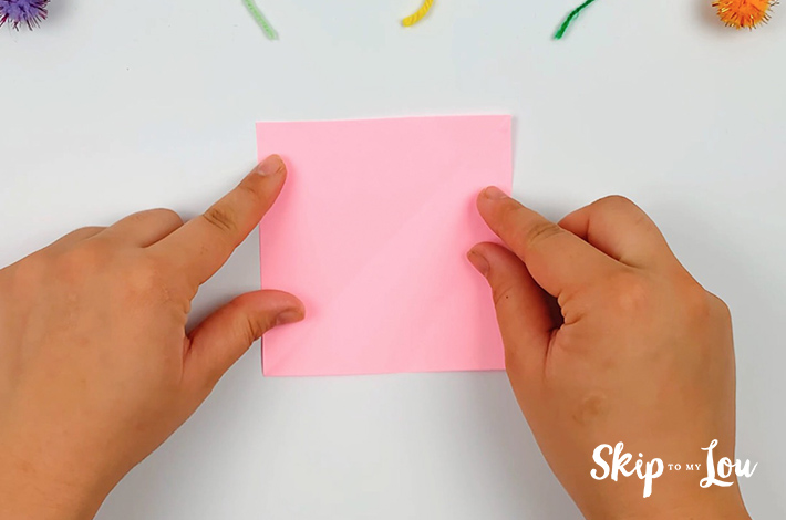 Two hands holding a square pink piece of paper.