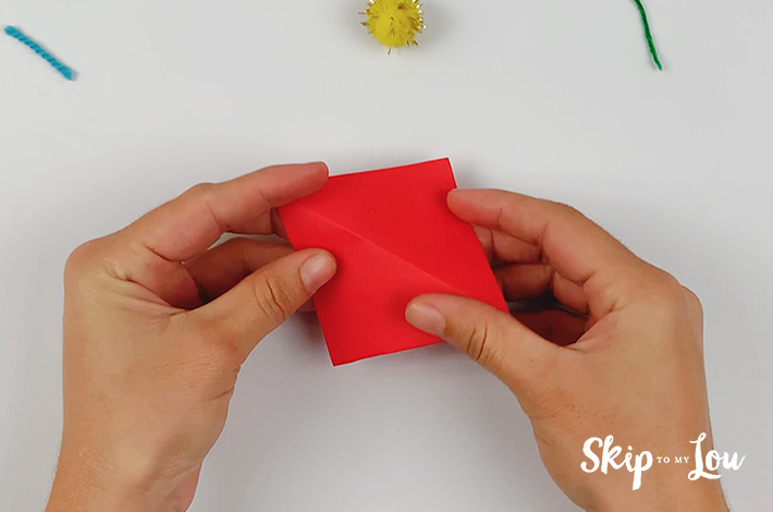 Two hand folding the red square origami paper to form two triangles.