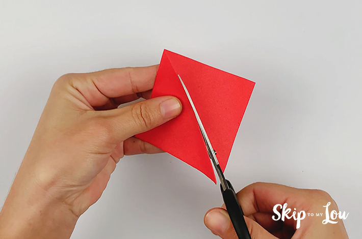 A pair of scissors cutting along the fold line in the red square piece of paper to form two triangles.