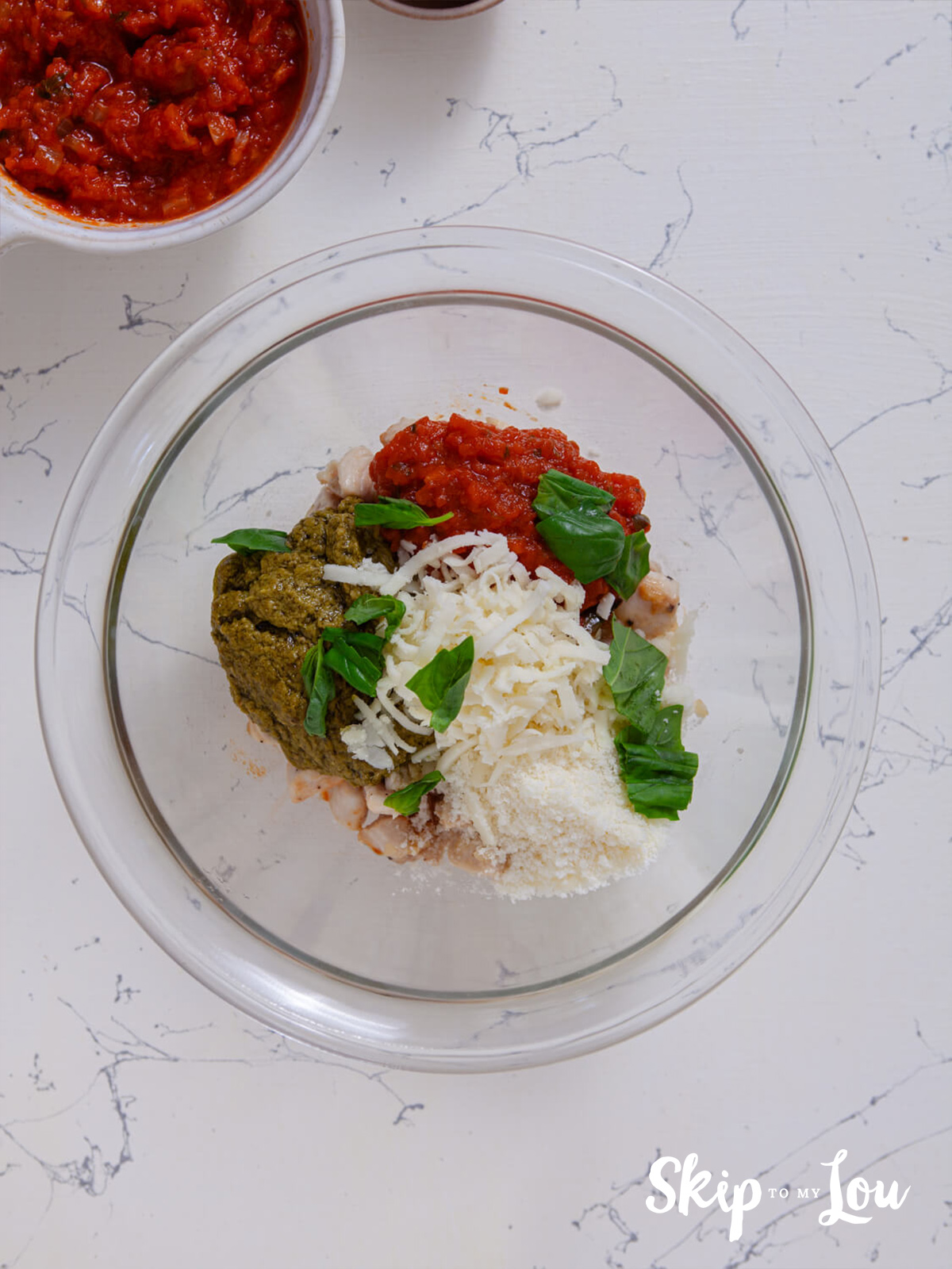 A large mixing bowl containing the cooked chicken, 1/4 cup of marinara sauce, pesto, mozzarella, parmesan, fresh basil and salt and pepper.