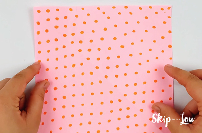 Two hands holding a pink piece of square paper with orange dots on it.