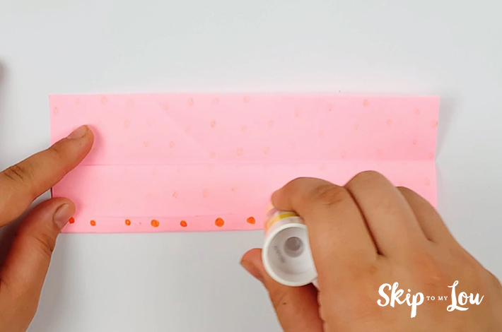 Two hand placing glue along the bottom of the unfolded pink paper with orange dots.
