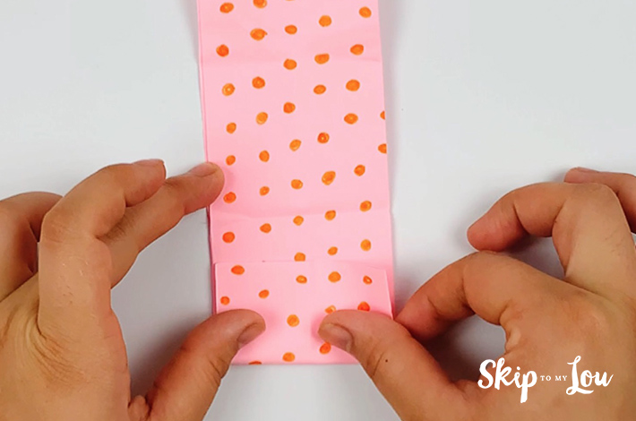 Two hands making a fold at the bottom of the pink paper with orange dots.