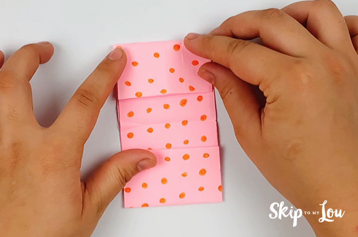 Two hands making a fold at the top of the pink paper with orange dots.