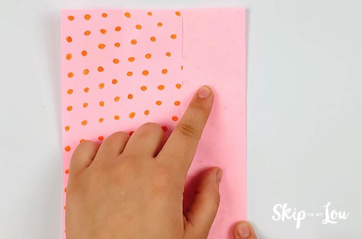 Two hands holding a pink piece of square paper with orange dots on it.