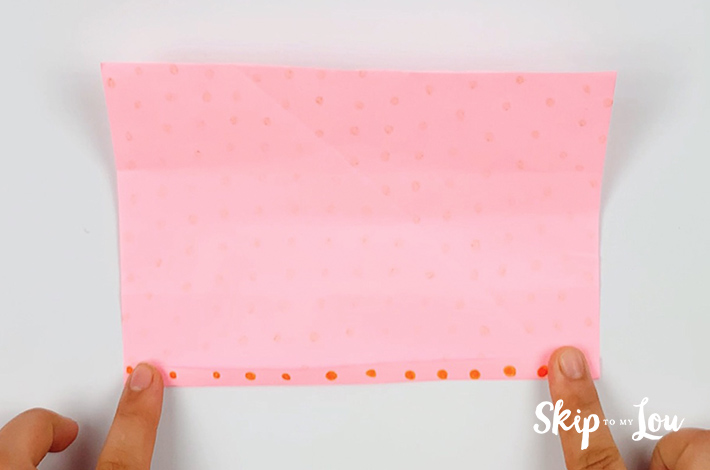 Two hands making a small fold at the bottom of the bigger piece of pink paper with orange dots.
