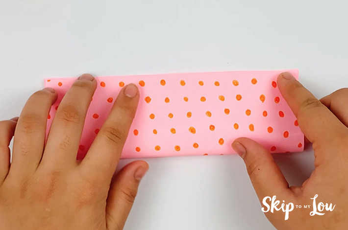Two hands folding the of pink paper with orange dots in half from the bottom to the top.
