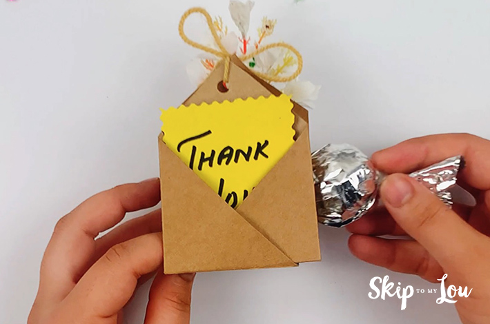 Two hands holding a completed party favor with a chocolate in the middle and a thank you note.