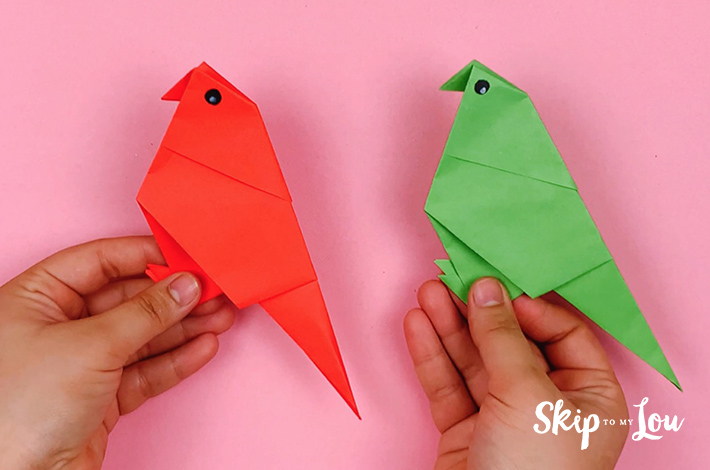 Two hands holding two compelted origami parrots, one red and one green.