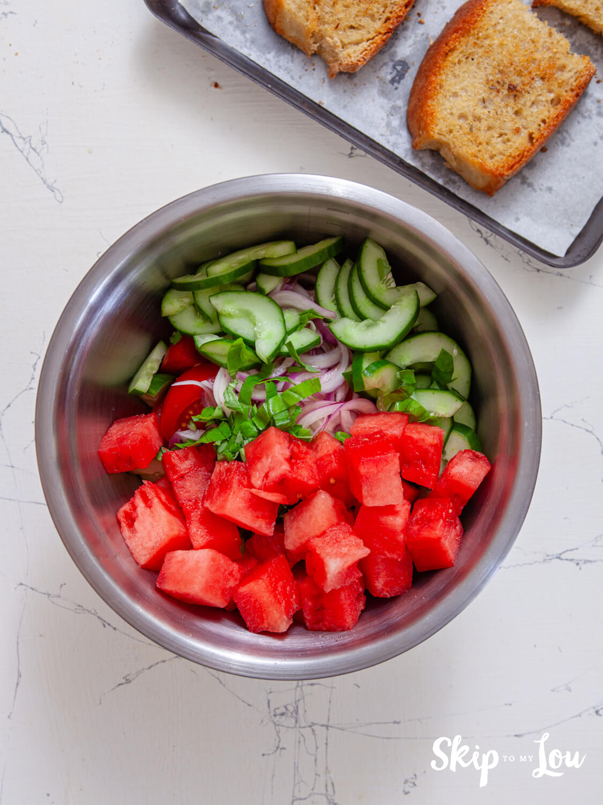 Watermelon, feta, basil, red onion and cucumber in a bowl, ready to be combined.