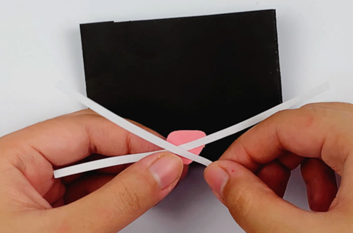 Two hands holding a smaller piece of paper for the cat's nose and longer strips of white paper for its whiskers.
