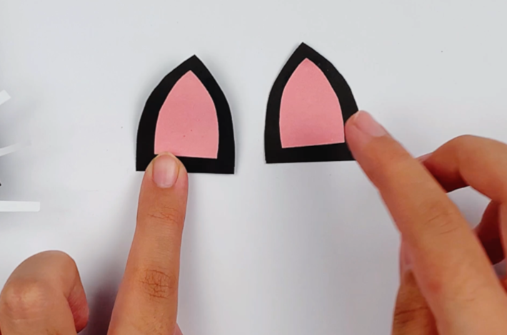 Two hands glueing two smaller pink ear shapes on top of the larger black ear shapes.