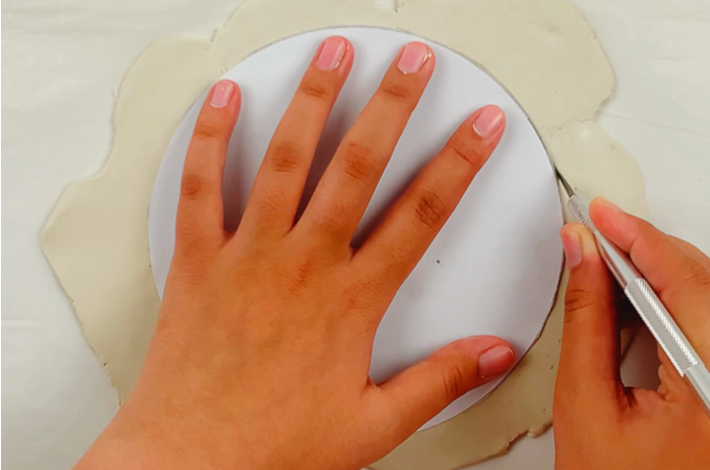 A hand holding the plate down while the other cuts the modeling clay around the edge of the plate. 