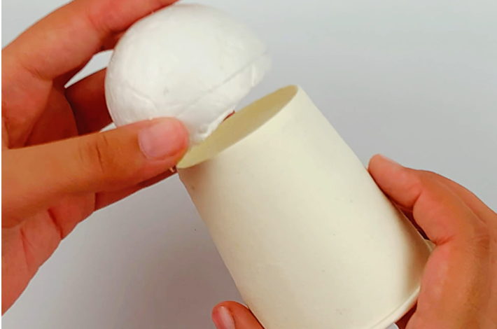 Two hands placing the styrofoam egg on the underside of the cup, which has been turned upside down. 
