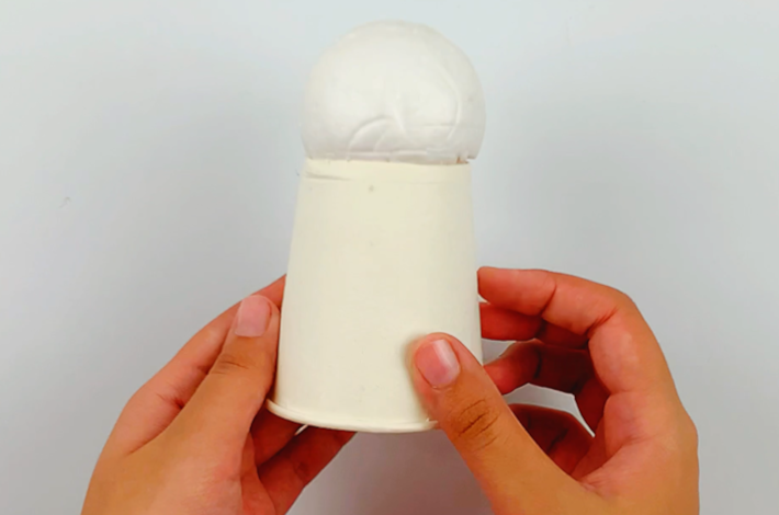 Two hands holding the cup with the stryrofoam egg on top of it. 