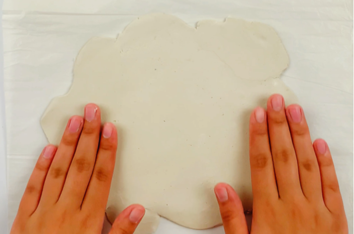 Two hands rolling the modeling clay out  as flat as possible while placed on baking paper. 