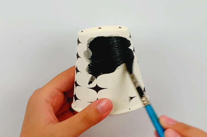 Two hands usinga  brush and black craft paint to paint the paper cup. 