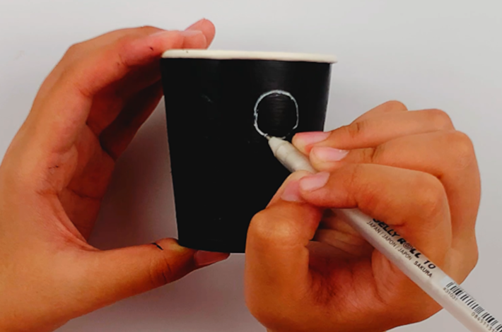 A hand holding the cup while the other draws eyes using a white marker. 