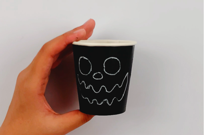 A hand holding up the paper cup with the eyes, nose and mouth drawn on it. 