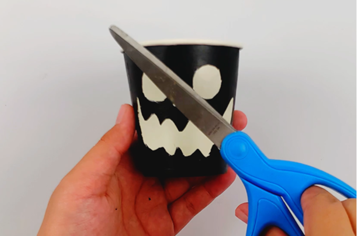 A hand holding scissors and the paper cup where the eyes, nose, and mouth outlines have been cut out. 