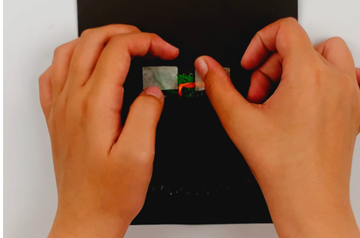 Two hands securing the orange thread on the back of the poster card and opposite the green thread using painter's tape. 