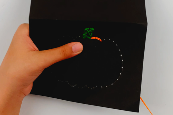 Two hands starting to thread the outline of the pumpkin using the needle and orange string.