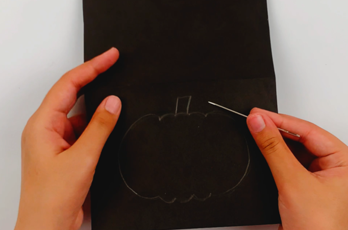 Two hands holding a needle with the poster card and the outline of the pumpkin in pencil below. 