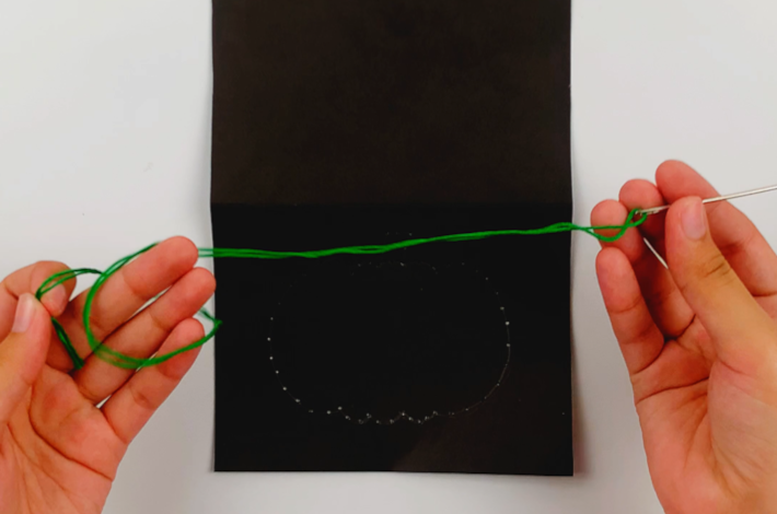 Two handles holding the needle above the A4 poster board with the green string threaded through it. 