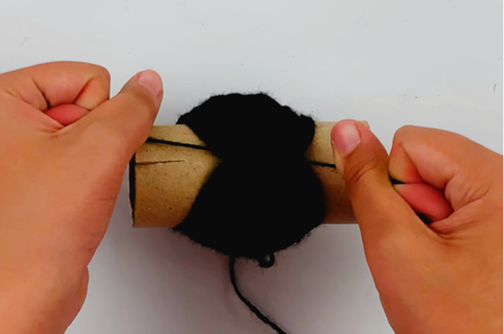 Two hands tying the loose ends of the wool around the wrapped wool to secure it.