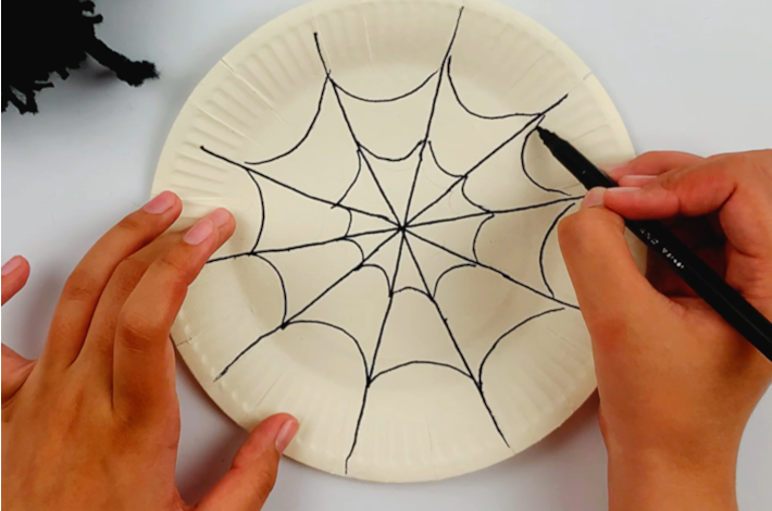 Two hands using a black marker to trace the spider web onto the paper plate.