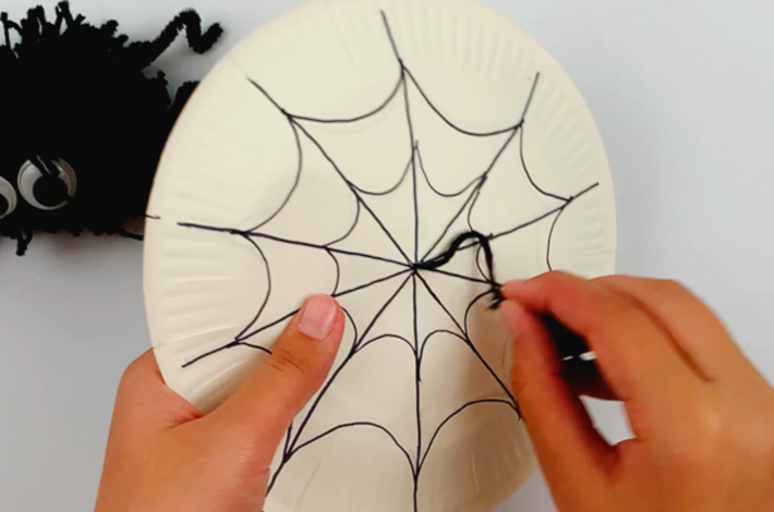 Two hands feeding the string from the pom pom spider through the hole in the paper plate.