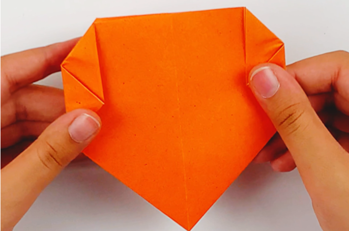 Two hands holding the orange paper after the same folds have been made on the left-hand side. 