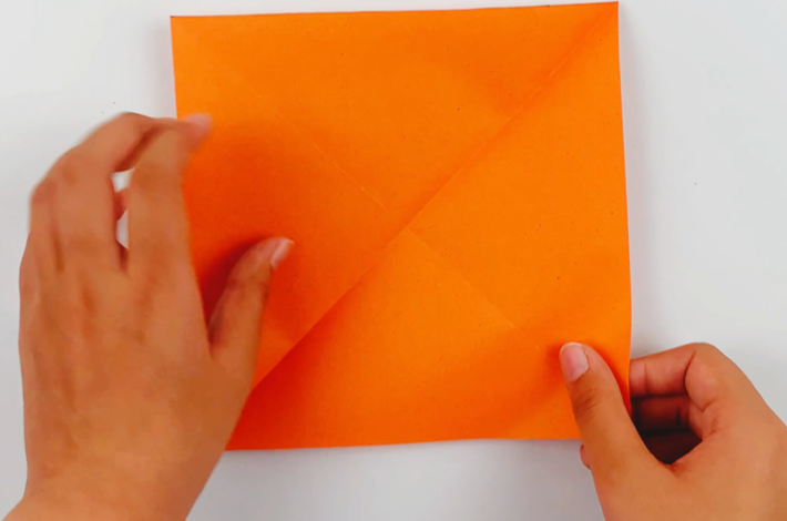 Two hands opening the orange paper to reveal an X-fold line from the two folds that have been made. 