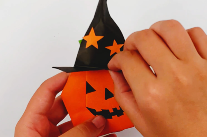 Two hands placing the finished witch's hat on the Halloween pumpkin.