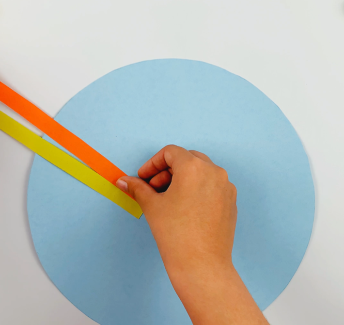Two hands gluing the strips of colored paper onto the blue paper circle. 