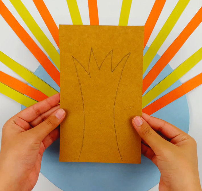 Two hands holding a brown piece of cardboard with a tree trunk shape drawn on it.