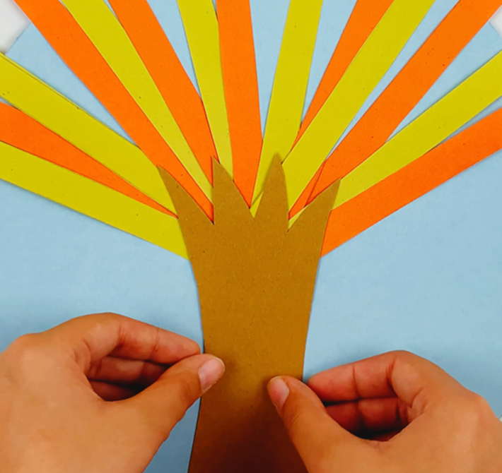 Two hands gluing the cut out tree trunk shape to the bottom of the blue paper. 