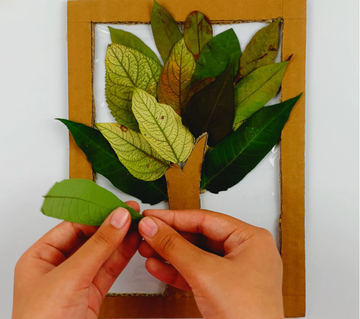 Two hands plaicing a final leaf on the fall tree to cover the whole area at the top of the frame. 