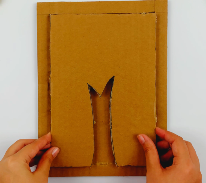 Two hands holding the inside section of the cardboard that has been cut out along the border and inside the tree stump. 