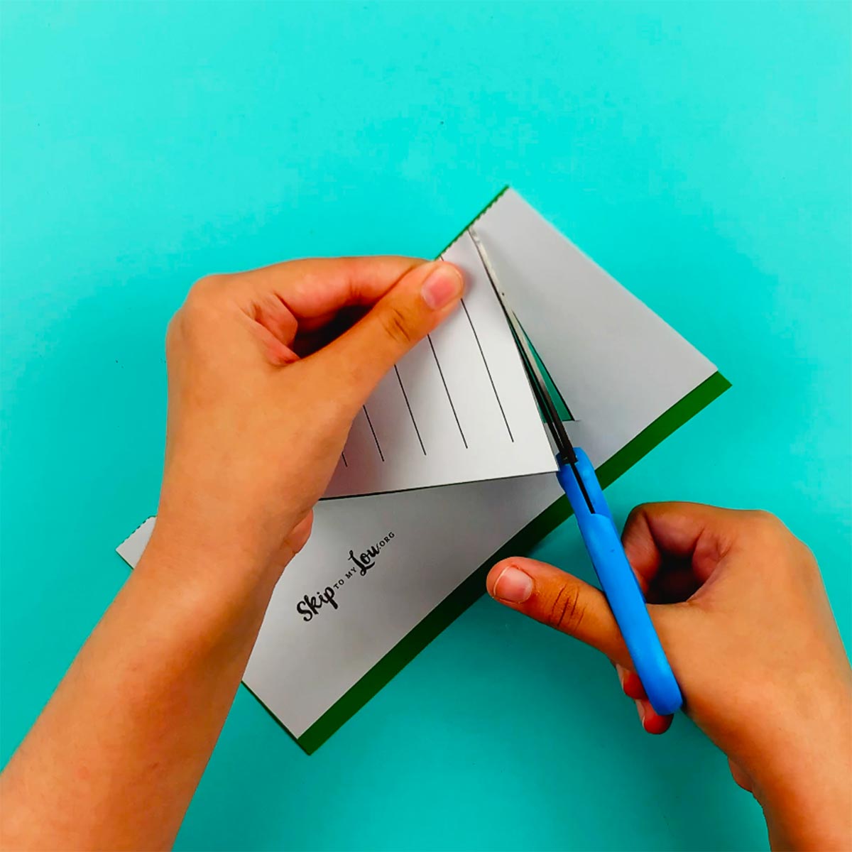Two hands placing the template over the green paper and cutting the outline of the christmas tree.