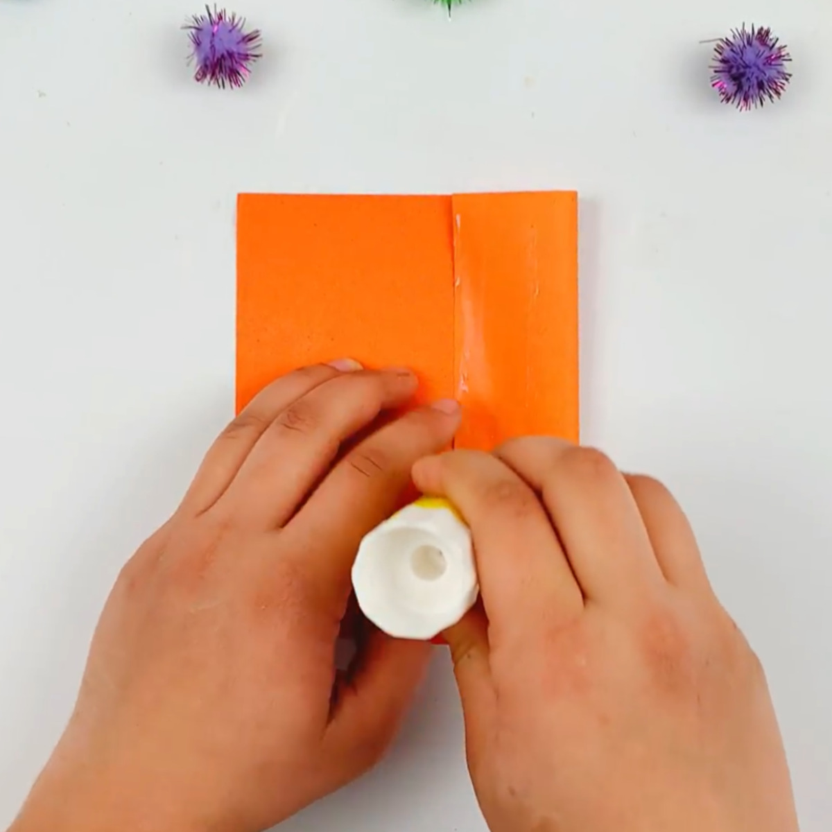 Two hands glueing down the right hand side of the orange paper after folding it over to the center.
