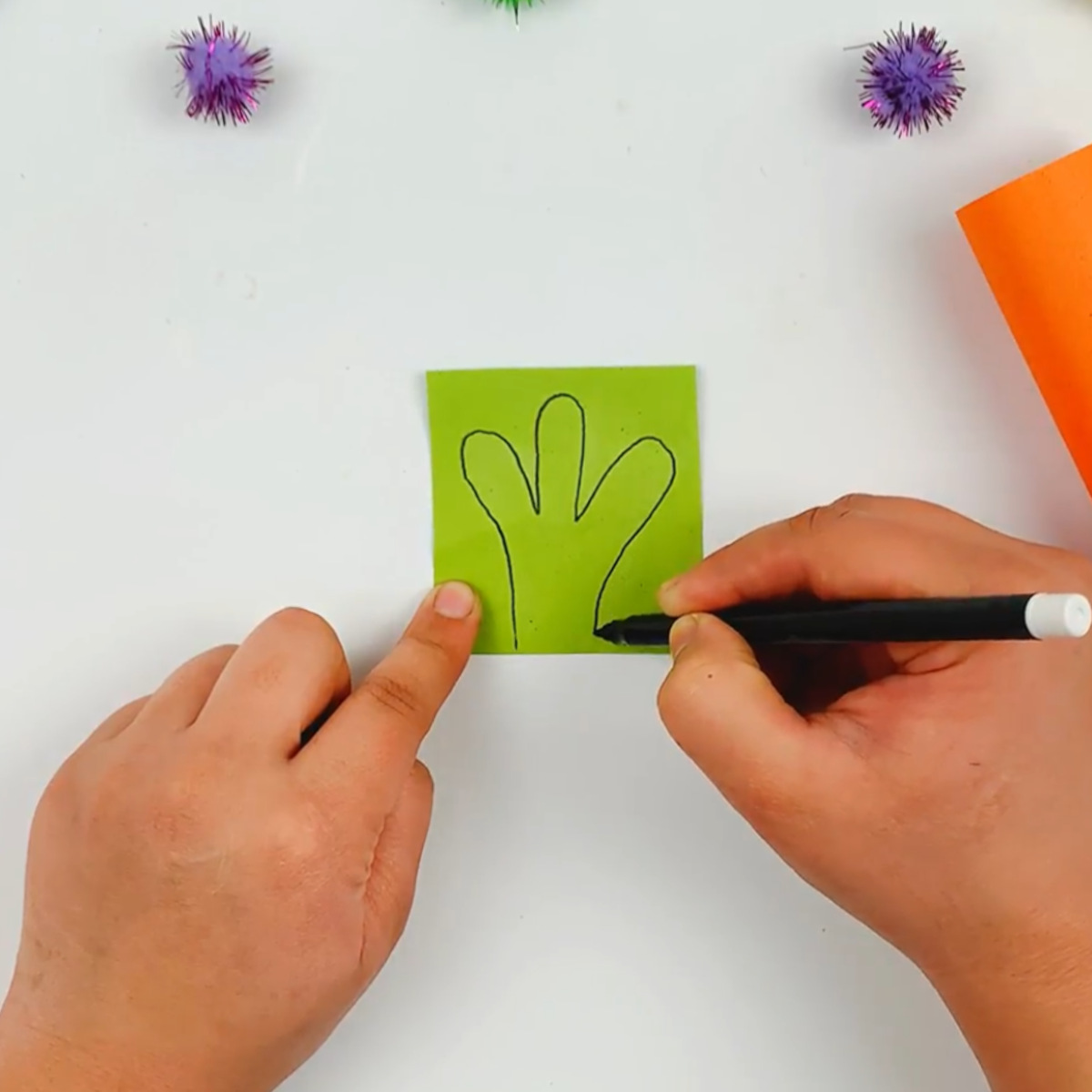 Two hands drawing the top section of the carrot on a piece of green paper.